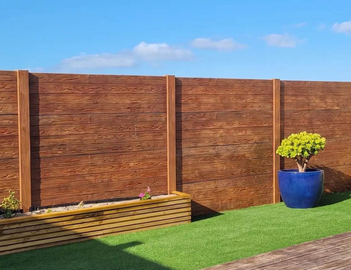 Muro pré-moldado em betão com textura de madeira em tom escuro, com canteiros e relvado num jardim
